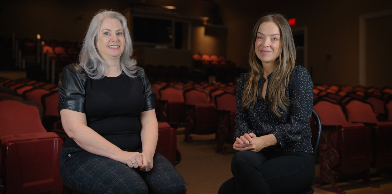 Two ladies sit in chairs in an empty theatre.